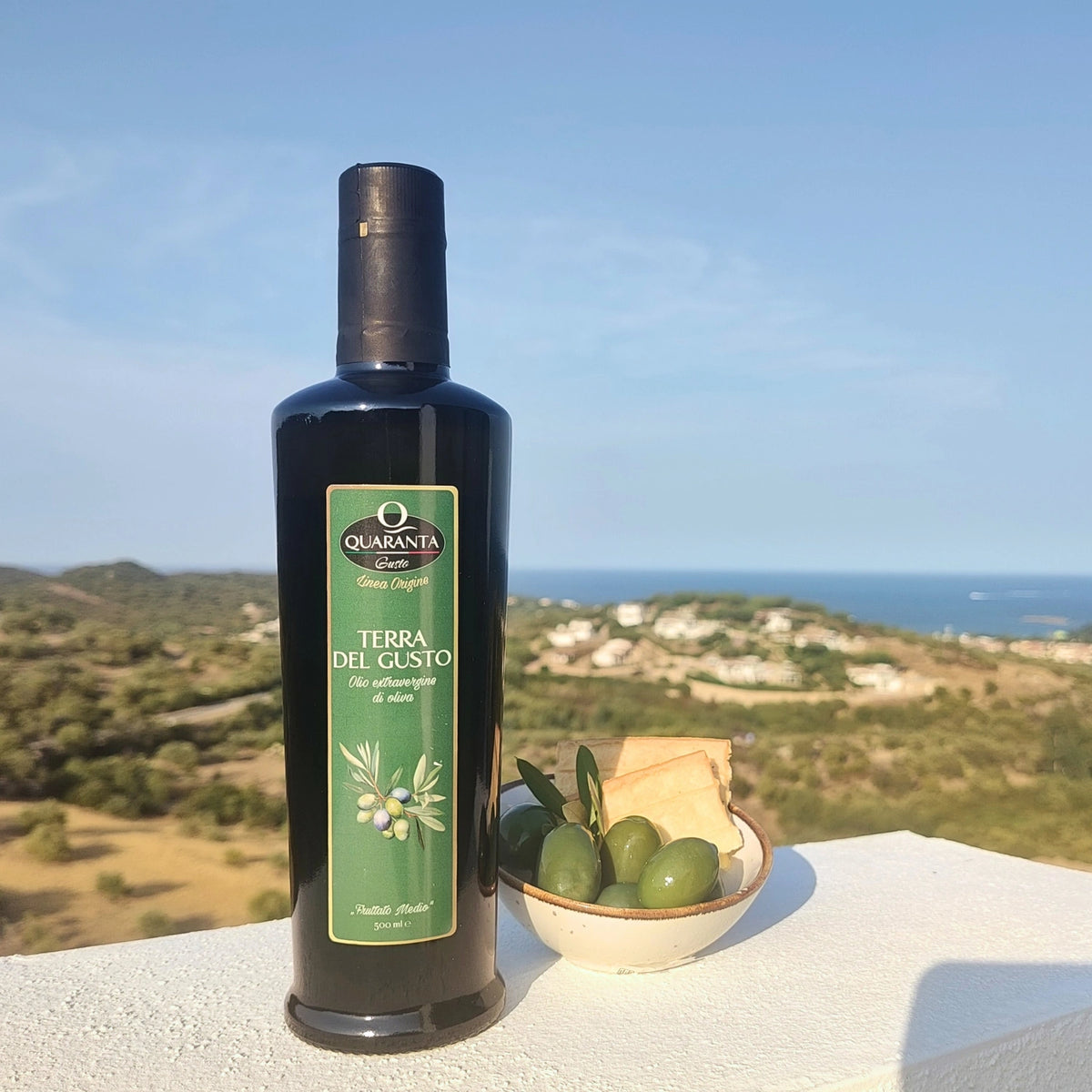 Quaranta Gusto Olivenöl-Flasche auf Sardinien; daneben grüne Oliven und Pane Carasau, im Hintergrund die Costa Smeralda.